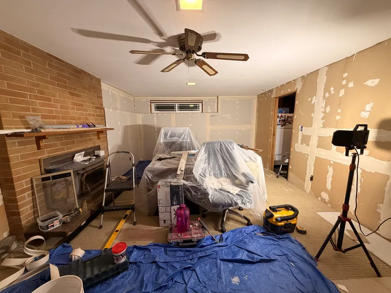 Room with the paneling removed, exposed patched drywall, a brick fireplace wall, plastic sheeting and a tripod