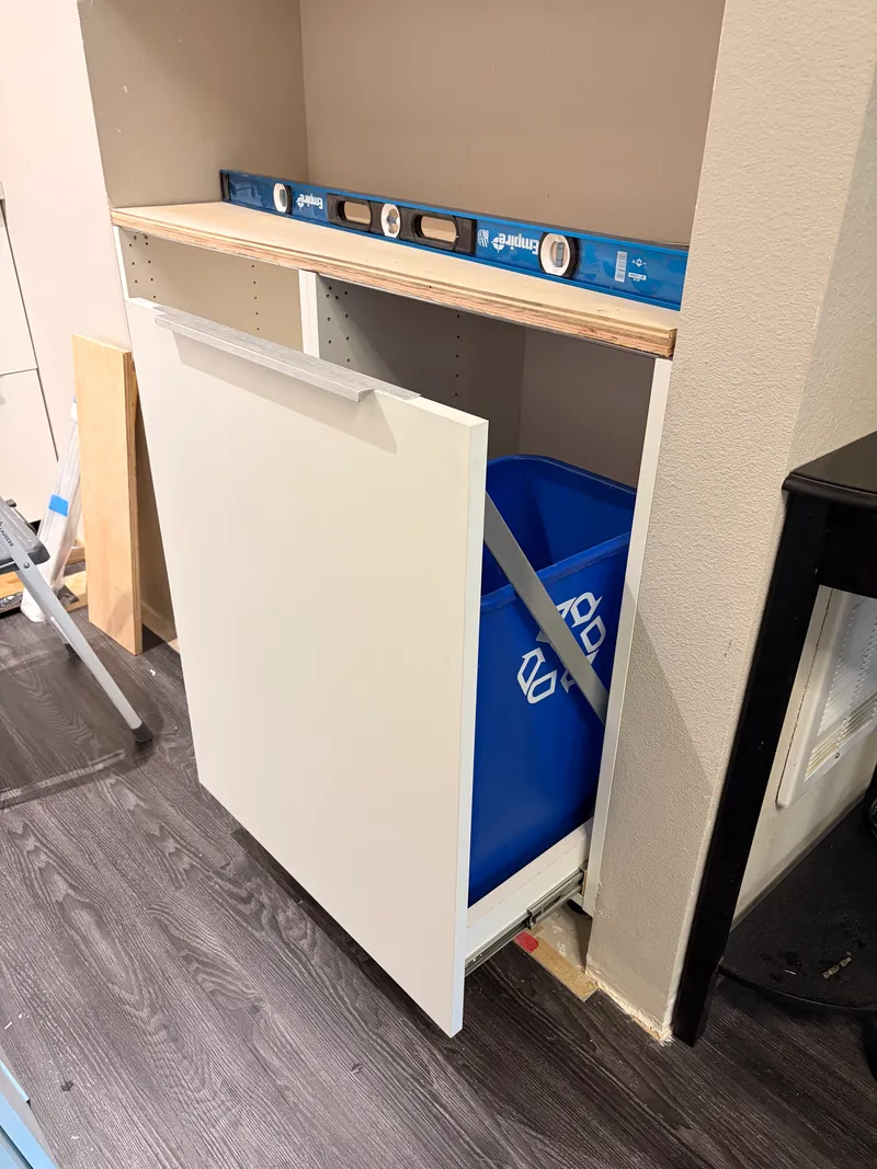 White cabinet door open on a custom pull-out trash bin, with a wood shelf above