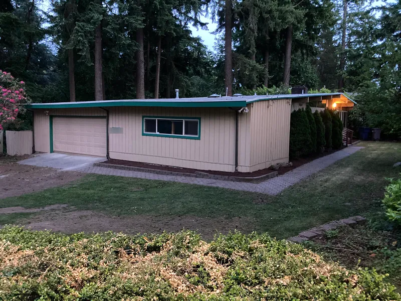 Single-story house with cream vertical siding, green trim and a garage, set among tall evergreen trees