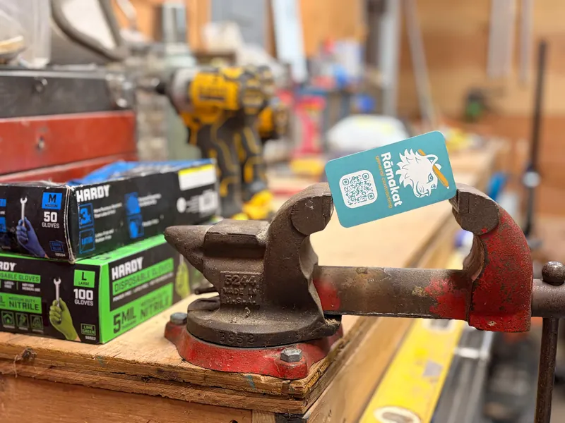 A pair of locking pliers on a workbench holding a Rāmakat tag, with tools and supplies behind