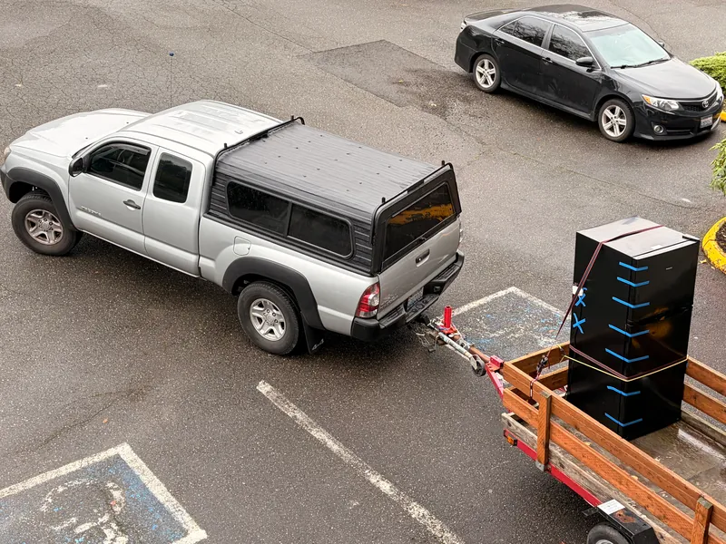 Silver Toyota Tacoma with a black canopy towing a red wood-sided trailer carrying a large black appliance